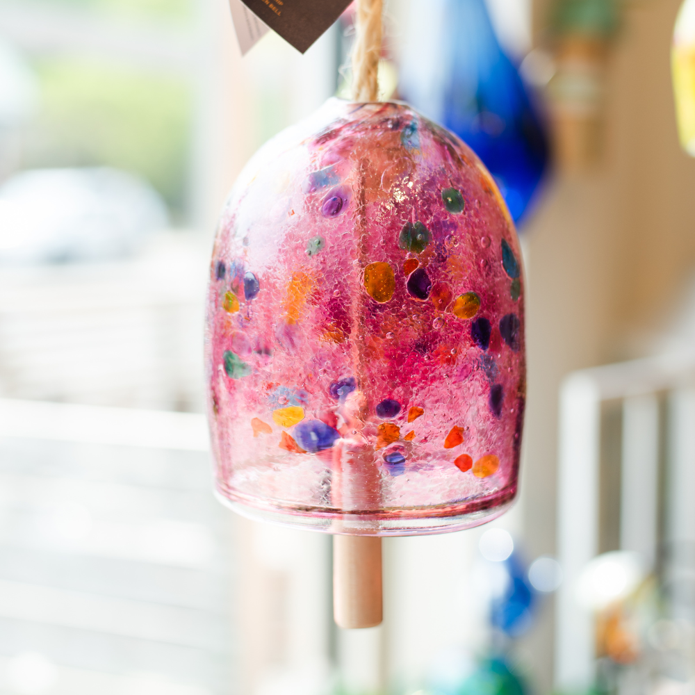 Pink bell with blue, purple, teal, orange, red, yellow and green spots with a rope and wood toggle in front of a window.