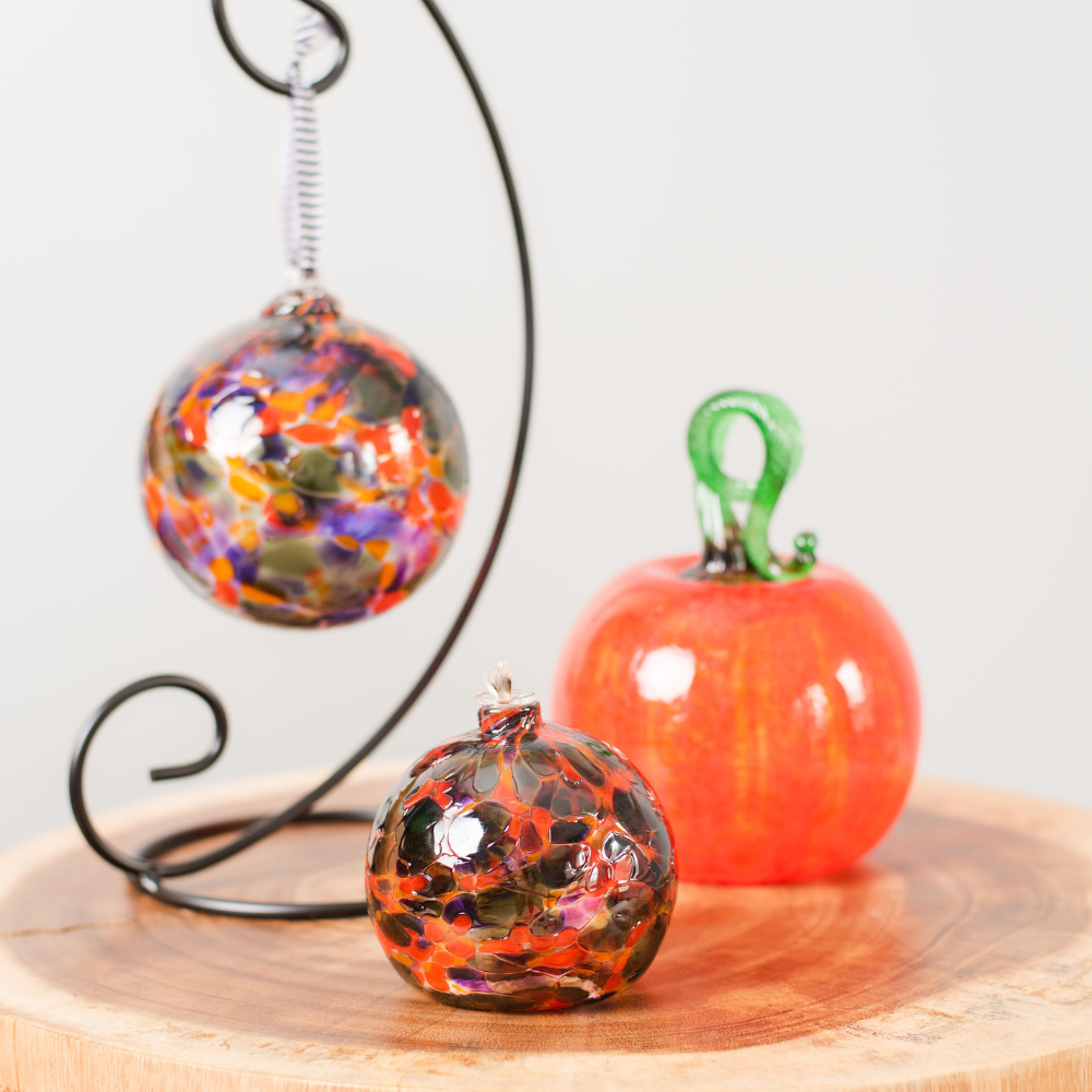 Round glass ball with flat base and wick with purple, orange and grey glass mosaiced around the entire surface on a wooden table in front of a round glass ball with purple, orange and grey glass mosaiced around the entire surface on a black curved ornament stand and a orange round pumpkin with a green stem