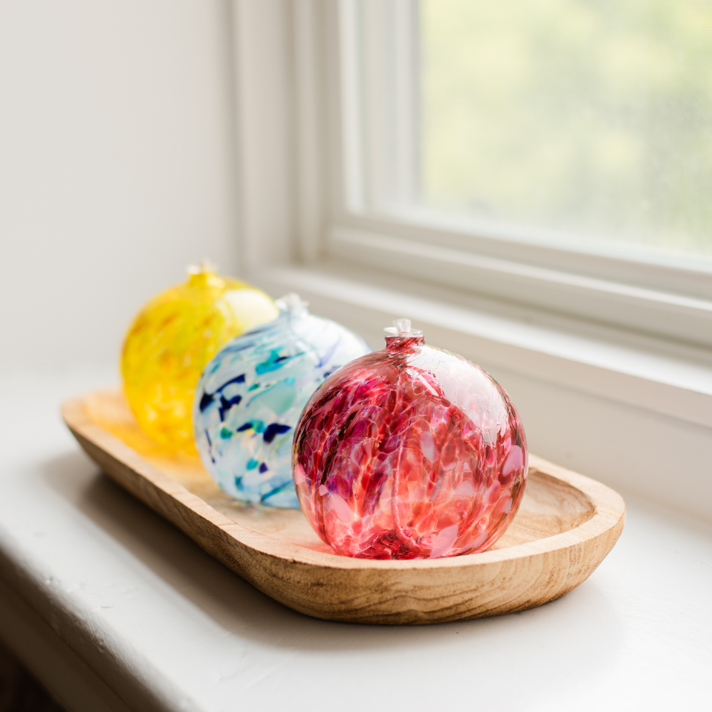 Pink round oil lamp, dark and light blue and white oil lamp and yellow oil lamp on a wood tray on a white window sill