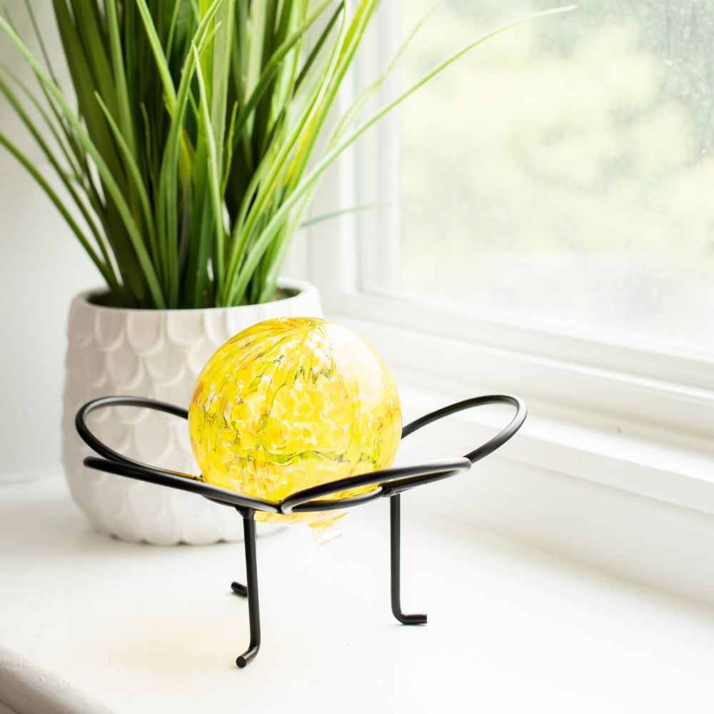 Yellow glass orb on a black flower garden holder on a white window sill with a green plant in the background