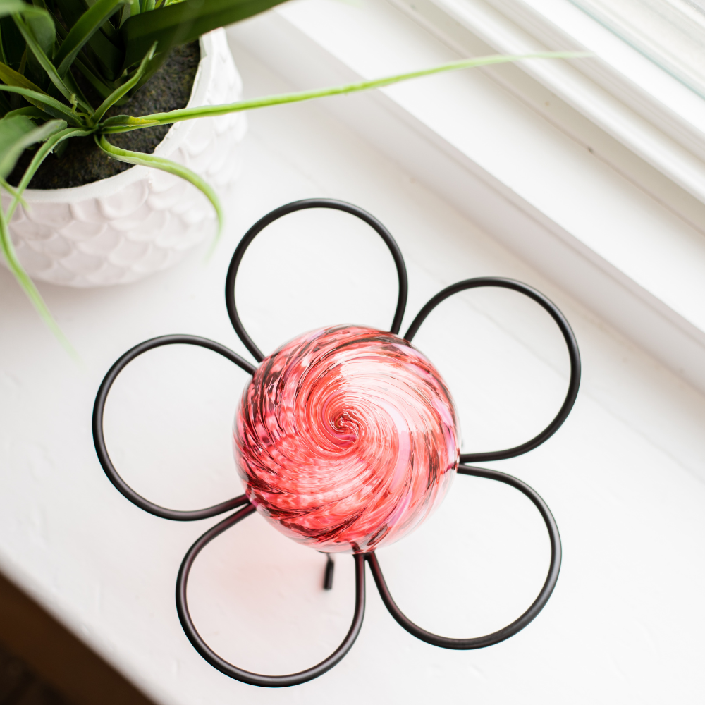 Pink glass orb, with a swirl on top, on a black flower garden holder, on a white window sill with a plant in the background