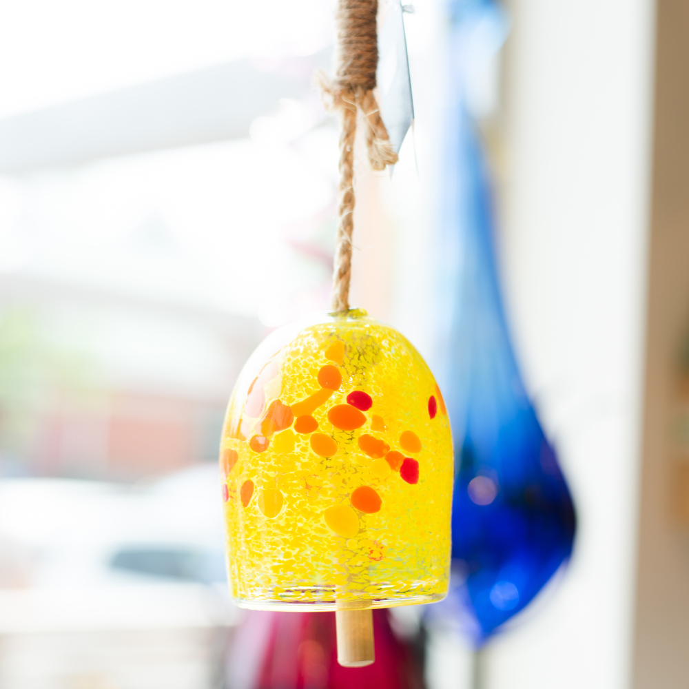 Opaque yellow bell with yellow, orange and red spots with a rope and wood toggle hanging in front of a window.