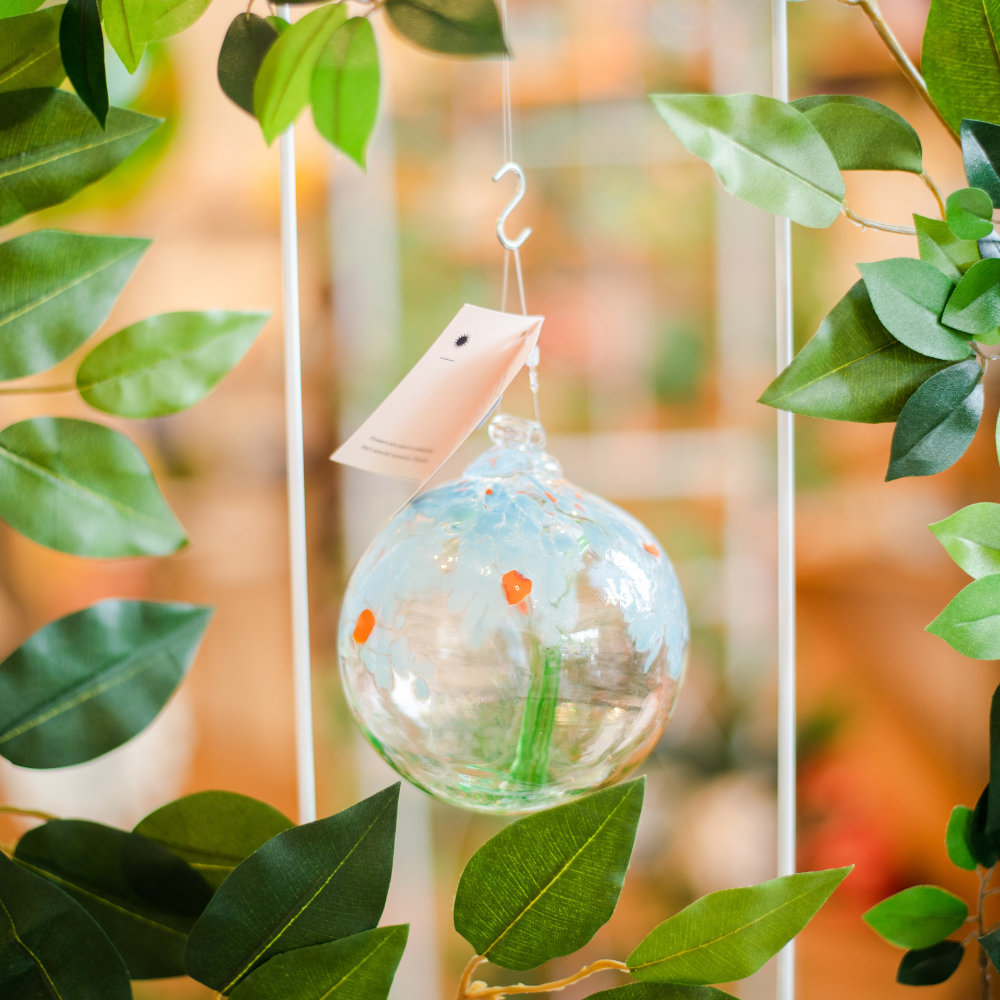 Clear glass orb with a green stem pulled from bottom to top to a canopy of blue opaque glass blossoms and orange centres hanging amongst greenery. 