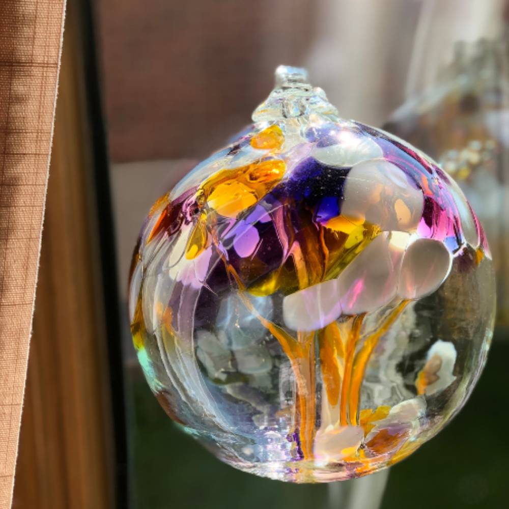 Clear glass orb with purple and pink glass pulled through the middle and yellow, white, purple and pink bits on top hanging in front of a window