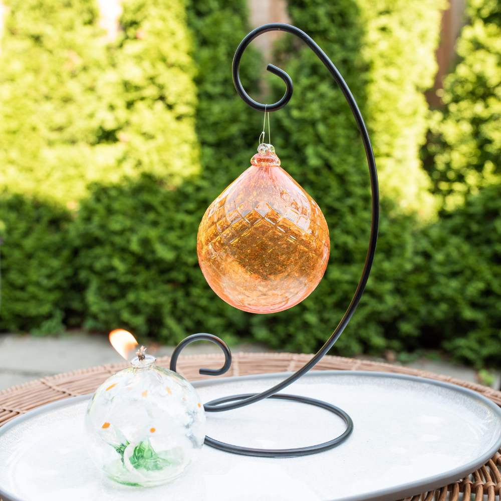 Lit White and green Friendship Blossom Oil Lamp and Orange Vibrant Fire Radiance Orb on a Large Curved Single Ornament Holder on a white plate with green trees in the background