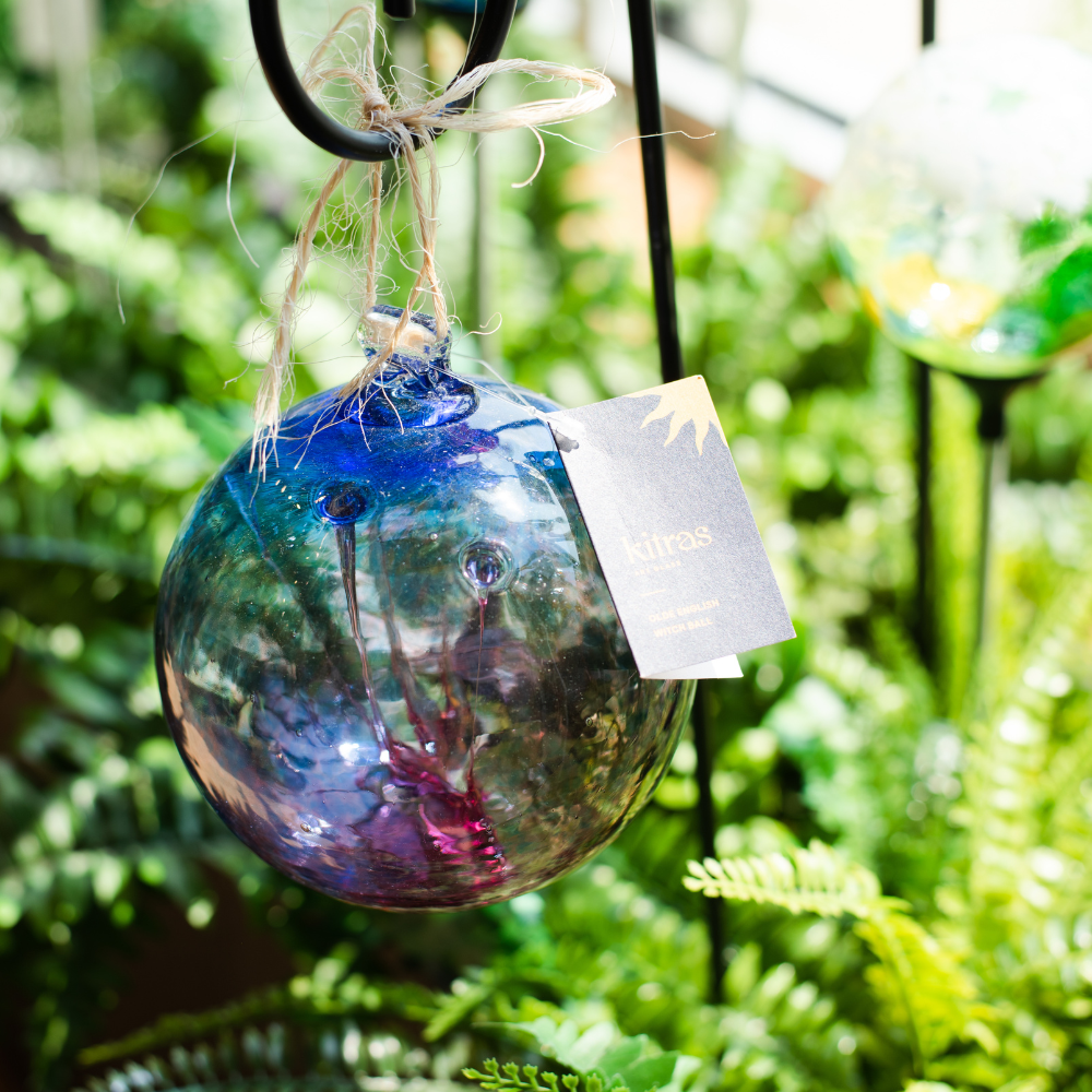 Glass orb with blue glass on top and pink glass on the bottom and webbing pulled from bottom to top on a black shepherd's hook stake in greenery.