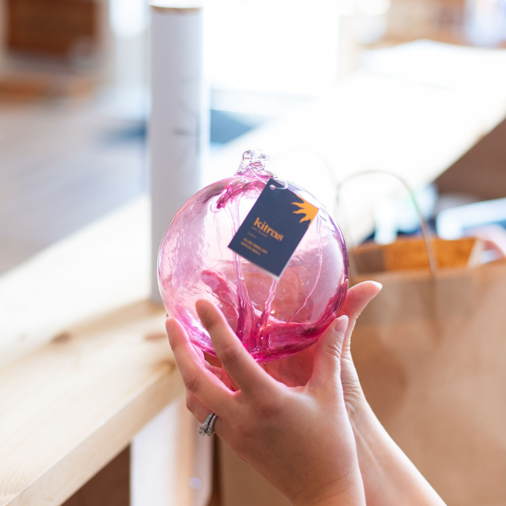 Pink glass orb with webbing pulled from bottom to top held in two hands in front of a gift back and wooden counter.