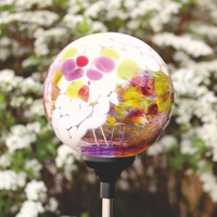 Glass orb with opaque white, yellow, lime and pink glass on the surface on a solar stake in a garden in front of flowers